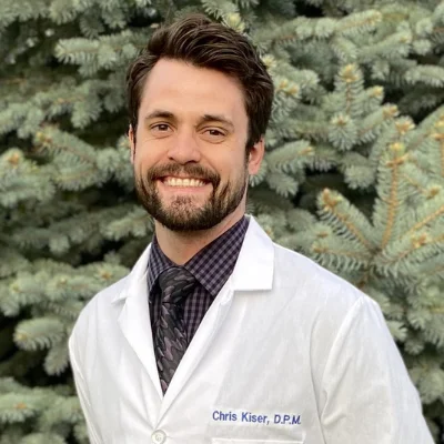 Dr. Chris Kiser, wearing white jacket and standing in front of pine tree.