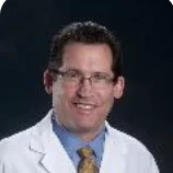 Headshot of Curtis Leviant, who has dark brown short hair, glasses and is wearing a Blue shirt, gold tie and white doctor's coat.