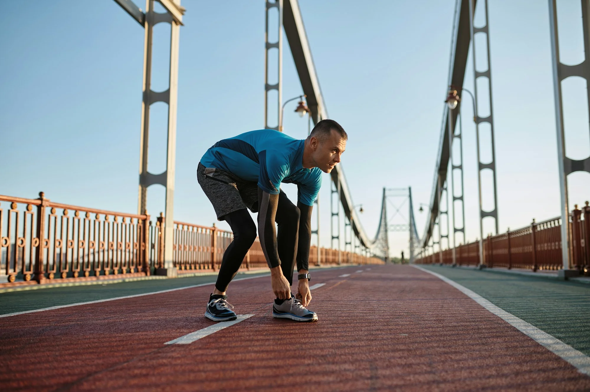 Young athlete man tying lace on running shoes during training on city bridge